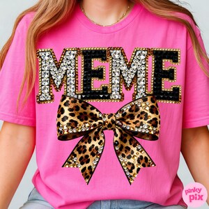 May include: A bright pink t-shirt with the word "MEME" in large, jeweled letters. Below the word is a large bow with a leopard print design, also adorned with jewels. The shirt is a vibrant and eye-catching piece.