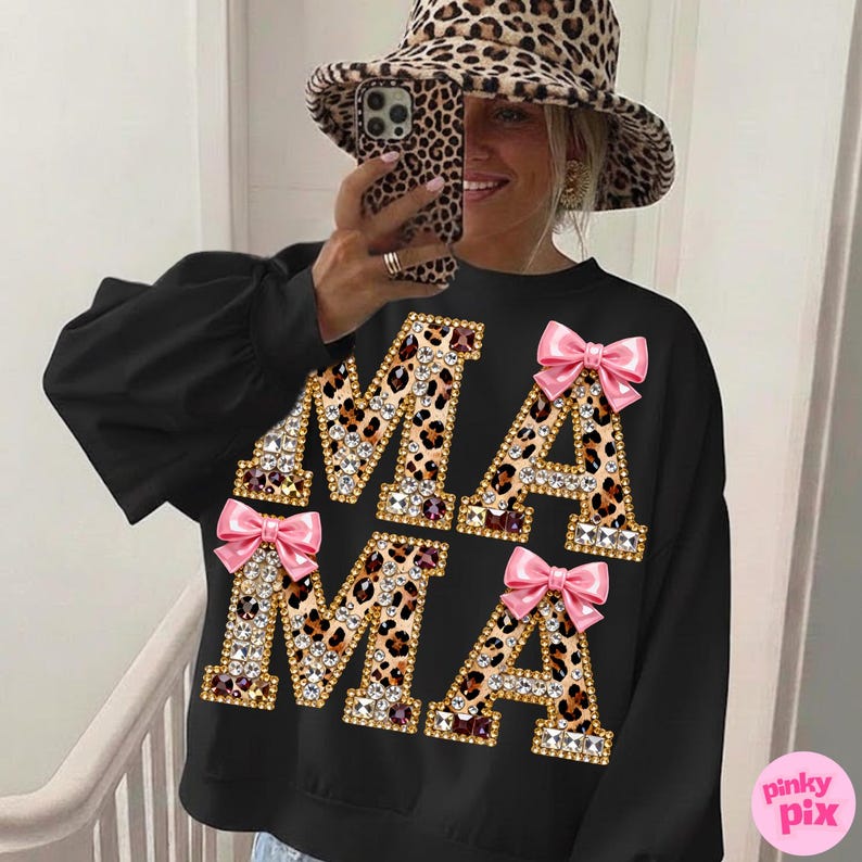 May include: Black sweatshirt featuring the word "MAMA" in leopard print letters, adorned with rhinestones and pink bows. The sweatshirt has long sleeves and a crew neck. A leopard print hat is also visible.