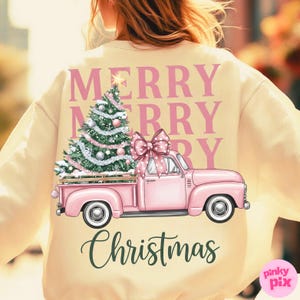 May include: Cream-colored sweatshirt with a pink Christmas truck graphic. The truck bed holds a decorated Christmas tree and is topped with a pink bow. The words "MERRY CHRISTMAS" are printed on the back.
