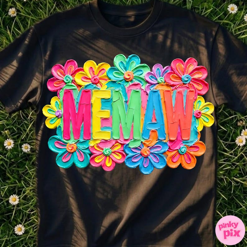 Puede incluir: Camiseta negra con la palabra "MEMAW" en letras coloridas y texturizadas, rodeada de vibrantes dise&ntilde;os florales multicolores. La camiseta se muestra sobre una superficie cubierta de hierba.