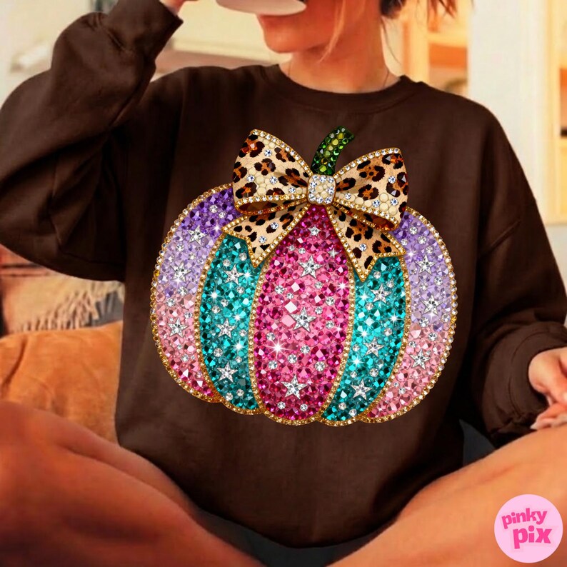 Op de afbeelding: Een donkerbruin sweatshirt met lange mouwen en een grote, centrale afbeelding van een pompoen versierd met veelkleurige, sprankelende, gefacetteerde edelstenen in roze, turquoise en paars, bekroond met een strik met luipaardprint en een groene steel.