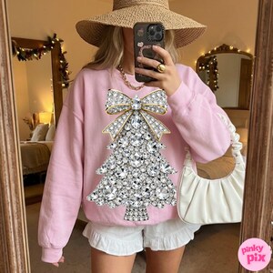 May include: A light pink sweatshirt featuring a sparkling Christmas tree design made of rhinestones and a gold bow. The sweatshirt is paired with white shorts. The image is taken in front of a mirror.