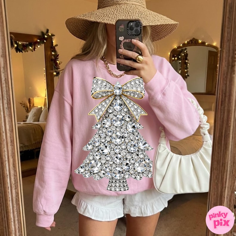May include: A light pink sweatshirt featuring a sparkling Christmas tree design made of rhinestones and a gold bow. The sweatshirt is paired with white shorts. The image is taken in front of a mirror.