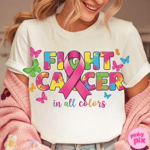 May include: White t-shirt with the words "FIGHT CANCER" in colorful, splattered letters, a pink ribbon, and butterflies. The phrase "in all colors" is written below. The shirt is part of a breast cancer awareness campaign.