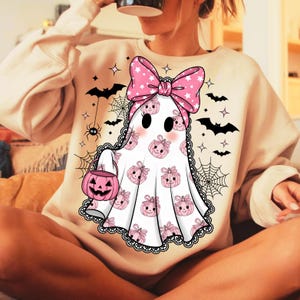 May include: A cream-coloured sweatshirt with a Halloween ghost design. The ghost is white with pink pumpkin and bow accents, holding a pink jack-o'-lantern. Black bats, spiders, and spiderwebs decorate the background, creating a festive, spooky theme.