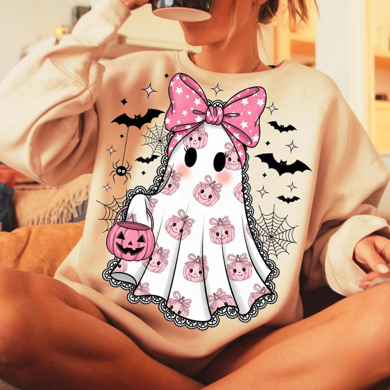 May include: A cream-coloured sweatshirt with a Halloween ghost design. The ghost is white with pink pumpkin and bow accents, holding a pink jack-o'-lantern. Black bats, spiders, and spiderwebs decorate the background, creating a festive, spooky theme.