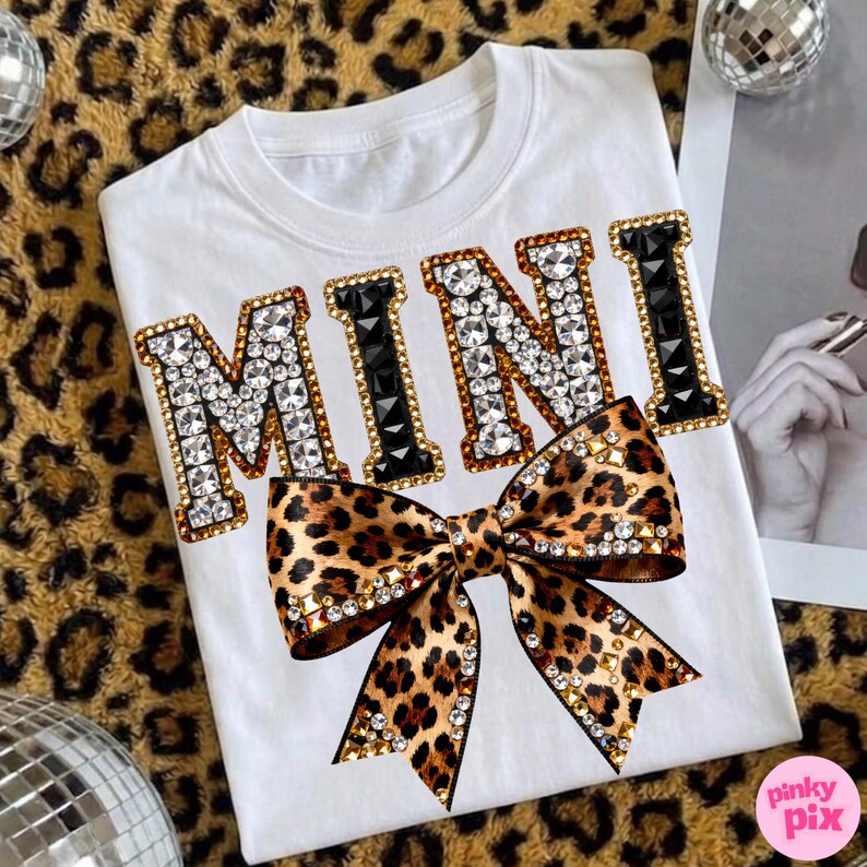 May include: White t-shirt with the word "MINI" in a jeweled design with black accents. A leopard print bow with rhinestone embellishments is below the word. The shirt is on a leopard print background.