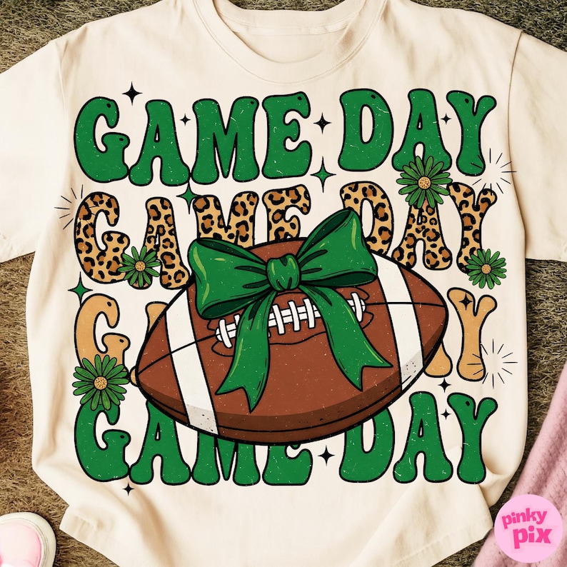 May include: Cream-colored t-shirt with a football and the words "GAME DAY" in green, leopard print, and tan. A green bow is tied around the football. The shirt has a retro, vintage design.