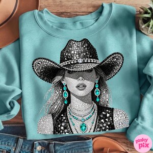 May include: A light blue sweatshirt featuring a rhinestone-studded illustration of a woman wearing a cowboy hat and turquoise jewelry. The hat and vest are embellished with rhinestones. The design is a striking blend of glamour and Western style.
