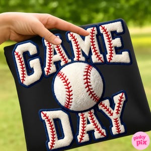 May include: Black baseball-themed item with the words "GAME DAY" in white, outlined in blue, and stitched with red thread. A baseball is in the center. The item is held by a hand.