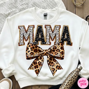May include: White sweatshirt with the word "MAMA" in jeweled letters, with a leopard print bow below. The letters are outlined in gold and filled with clear and black gems. The bow has gold accents.