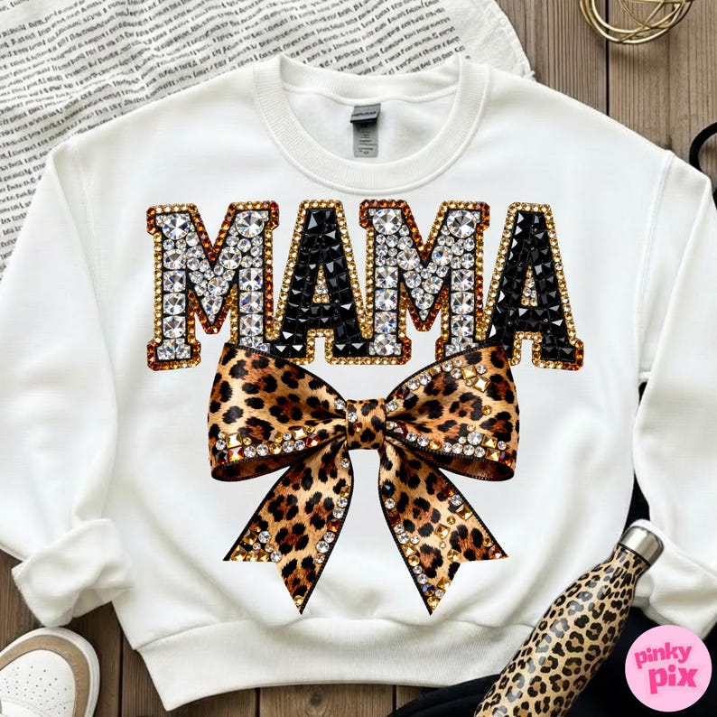 May include: White sweatshirt with the word "MAMA" in jeweled letters, with a leopard print bow below. The letters are outlined in gold and filled with clear and black gems. The bow has gold accents.