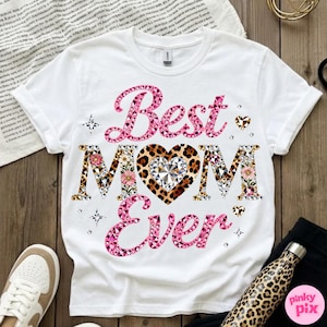 May include: White t-shirt with the text "Best MOM Ever" in pink and patterned letters. The heart in "MOM" has a leopard print design with a diamond center. Floral and geometric patterns decorate the letters.