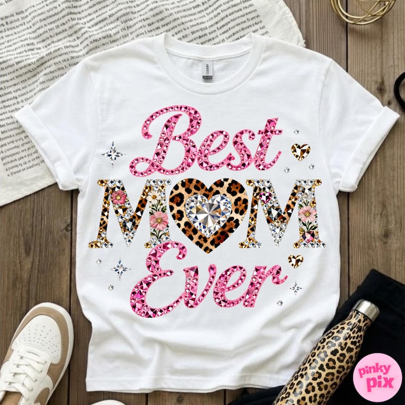 May include: White t-shirt with the text "Best MOM Ever" in pink and patterned letters. The heart in "MOM" has a leopard print design with a diamond center. Floral and geometric patterns decorate the letters.