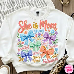 K&ouml;nnte beinhalten: Wei&szlig;es Sweatshirt mit dem Text "She is Mom" in rosa und orangefarbener Schrift. Das Design umfasst bunte Schleifen und W&ouml;rter wie "Strong", "Beautiful" und "Victorious". Eine Wasserflasche mit Leopardenmuster ist sichtbar.