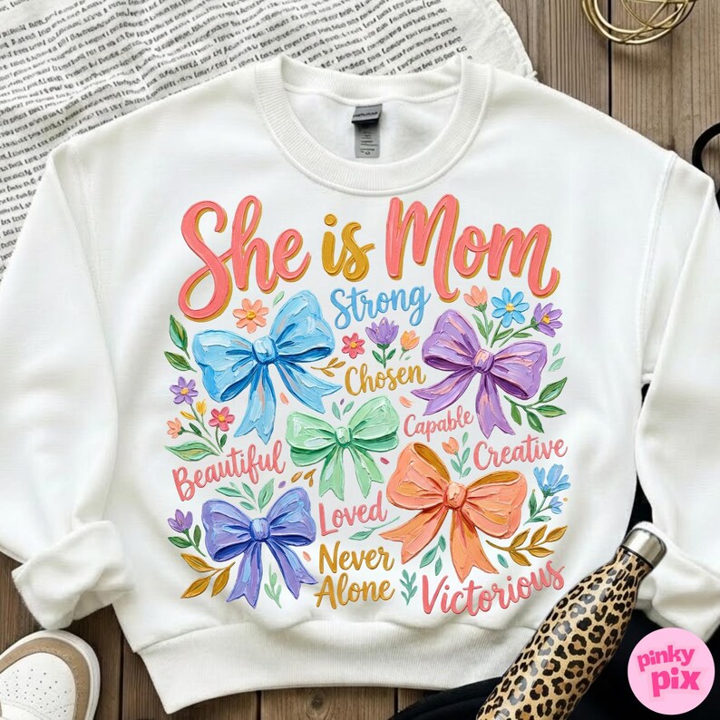 K&ouml;nnte beinhalten: Wei&szlig;es Sweatshirt mit dem Text "She is Mom" in rosa und orangefarbener Schrift. Das Design umfasst bunte Schleifen und W&ouml;rter wie "Strong", "Beautiful" und "Victorious". Eine Wasserflasche mit Leopardenmuster ist sichtbar.