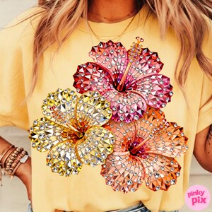 May include: Yellow t-shirt featuring a vibrant design of three hibiscus flowers. The flowers are embellished with sparkling crystals in shades of red, pink, yellow, and orange. The shirt is a solid yellow color.
