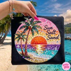 May include: Black tote bag featuring a vibrant, circular design with a sunset, palm trees, and the words "Sun Salt Sand." The design incorporates sparkling embellishments in various colors, including orange, pink, and blue, creating a beachy theme.