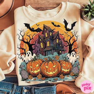 May include: Cream-colored sweatshirt featuring a Halloween-themed graphic. The design includes a haunted house, jack-o'-lanterns, ghosts, bats, and bare trees against a sunset backdrop. The graphic is in shades of orange, purple, and black.