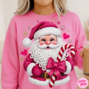 Puede incluir: Sudadera rosa con un diseño detallado de Papá Noel. El diseño incluye barba blanca, traje rojo y bastón de caramelo. El diseño incluye un lazo rosa, corazones, estrellas y adornos.