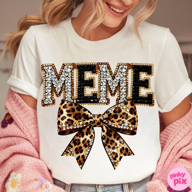 May include: White t-shirt with the word "MEME" in a jeweled design with black accents. Below the text is a leopard print bow with rhinestone embellishments. The shirt is a casual style.