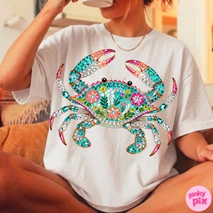 May include: White t-shirt featuring a detailed crab design. The crab is adorned with turquoise, pink, and clear crystal-like embellishments, with floral accents on its body. The design is centered on the shirt.