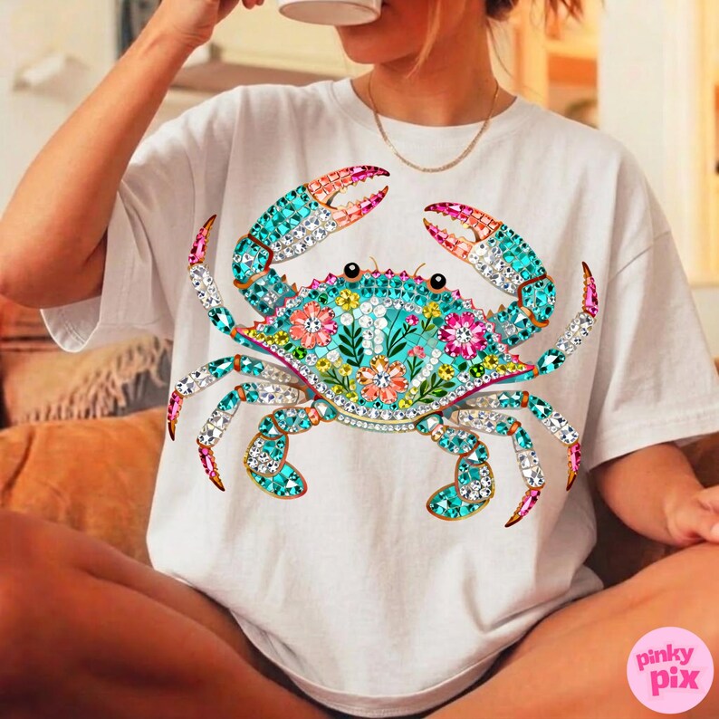 May include: White t-shirt featuring a detailed crab design. The crab is adorned with turquoise, pink, and clear crystal-like embellishments, with floral accents on its body. The design is centered on the shirt.