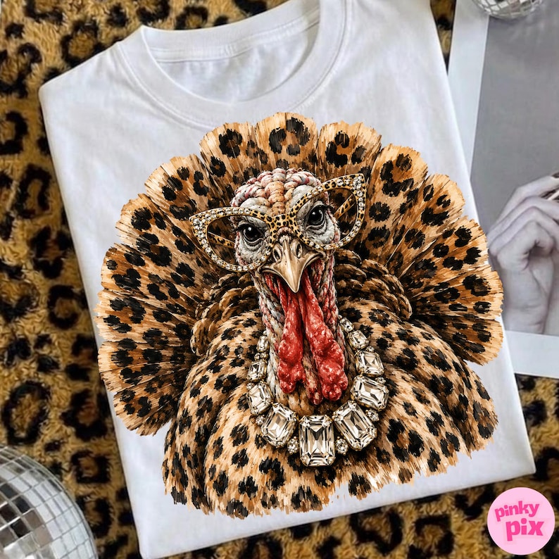 May include: A white t-shirt featuring a graphic print of a turkey with a leopard print pattern on its feathers. The turkey wears leopard print cat-eye glasses and a large, clear crystal statement necklace around its red, textured neck.