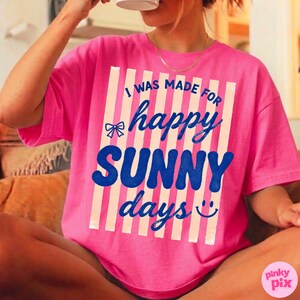May include: A bright pink t-shirt with the text "I WAS MADE FOR happy SUNNY days" in blue and white. The design includes a bow and a smiling face. The shirt has a relaxed fit.