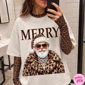 以下が含まれることがあります： クリーム色の半袖Tシャツ。茶色の大きな文字で「MERRY」と書かれ、その下にヒョウ柄の服と帽子、サングラスを身につけたサンタクロースのグラフィックが描かれています。ヒョウ柄の長袖シャツの上に重ね着されています。