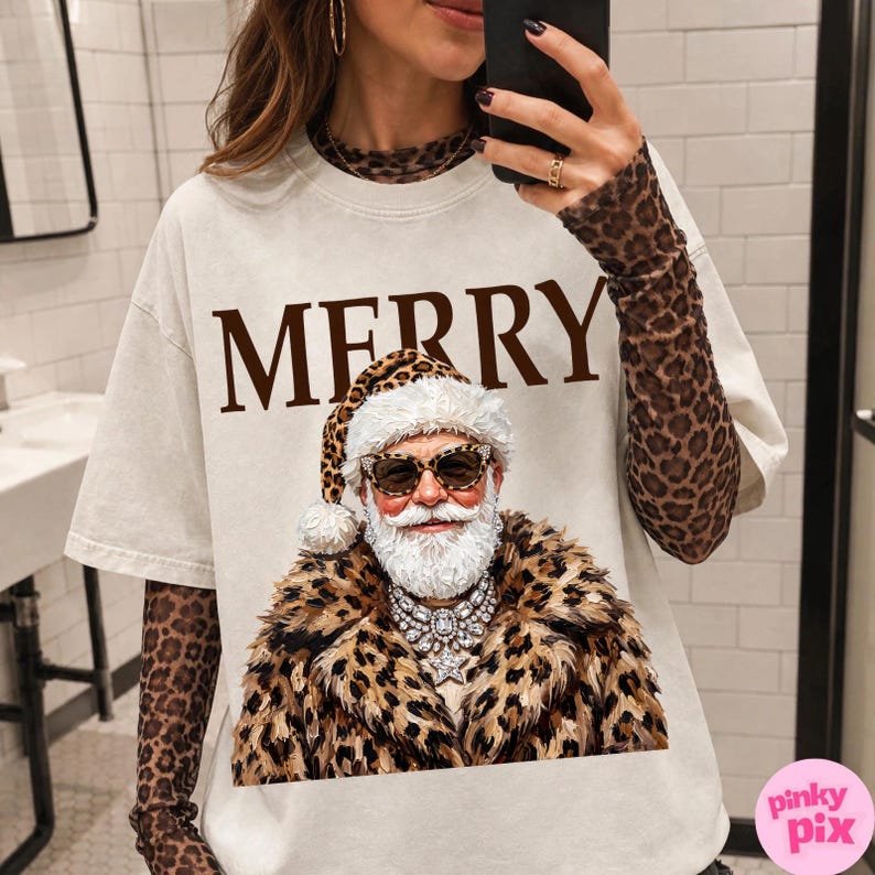 以下が含まれることがあります： クリーム色の半袖Tシャツ。茶色の大きな文字で「MERRY」と書かれ、その下にヒョウ柄の服と帽子、サングラスを身につけたサンタクロースのグラフィックが描かれています。ヒョウ柄の長袖シャツの上に重ね着されています。