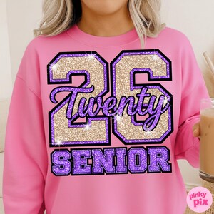May include: A pink sweatshirt with a glittery gold and purple graphic that reads "26 Twenty SENIOR". The number 26 is outlined in black and filled with gold glitter. The word "Twenty" is in a cursive font, and "SENIOR" is in block letters.