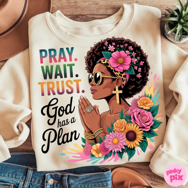 May include: Cream-colored sweatshirt with a graphic of a woman wearing sunglasses, a cross earring, and floral accents. The text "PRAY. WAIT. TRUST. God has a Plan" is printed in colorful letters. The design includes pink, yellow, and green hues.