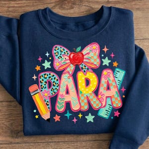May include: Navy blue sweatshirt with a colorful graphic design. The design includes the word "PARA" in large, decorated letters, a bow with an apple, a pencil, a ruler, and stars. The sweatshirt is folded.