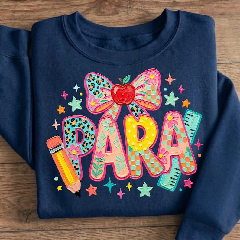 May include: Navy blue sweatshirt with a colorful graphic design. The design includes the word "PARA" in large, decorated letters, a bow with an apple, a pencil, a ruler, and stars. The sweatshirt is folded.
