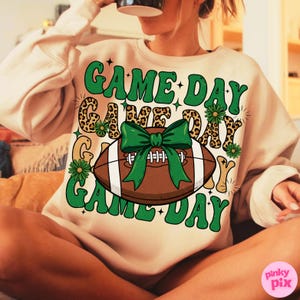 May include: A beige sweatshirt with a football graphic and the words "GAME DAY" in green. The football has a green bow and white laces. The design includes leopard print accents and floral details, perfect for game day.