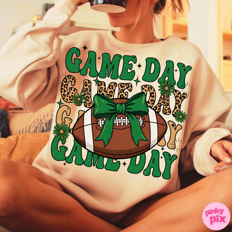 May include: A beige sweatshirt with a football graphic and the words "GAME DAY" in green. The football has a green bow and white laces. The design includes leopard print accents and floral details, perfect for game day.
