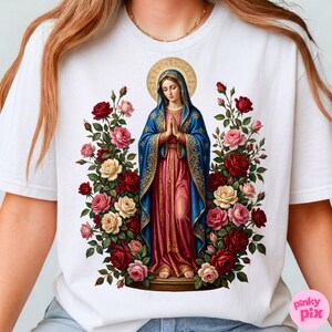 May include: White t-shirt featuring a religious illustration of a woman in a blue and pink robe, surrounded by roses in shades of red, pink, and cream. The woman has a gold halo and is in a prayerful pose.