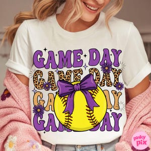 May include: White t-shirt with a softball graphic. The design features the words "GAME DAY" in purple, leopard print, and tan, with a purple bow and a yellow softball. The shirt has a crew neck and short sleeves.