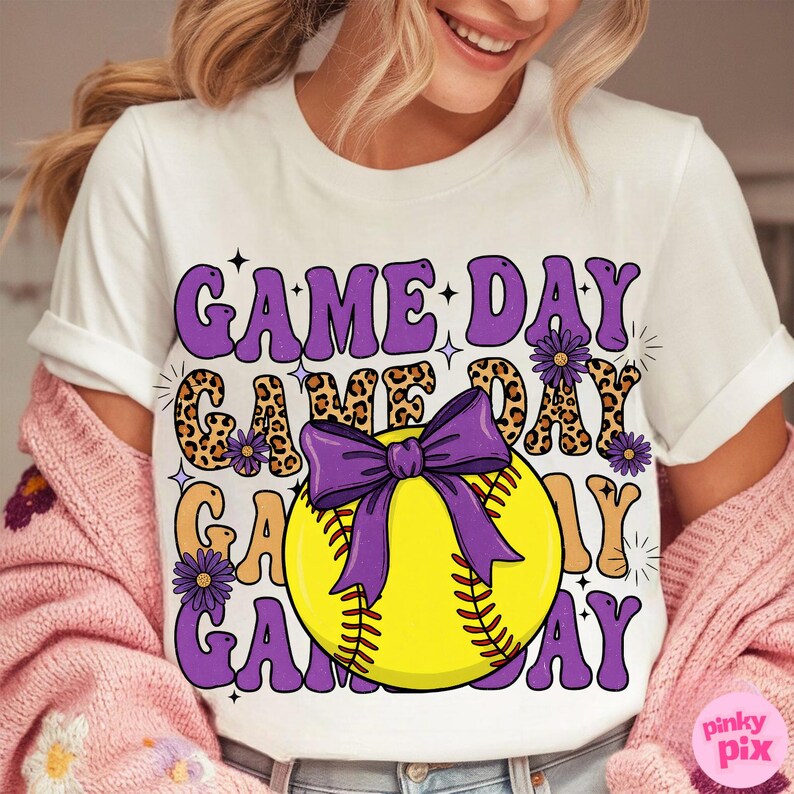 May include: White t-shirt with a softball graphic. The design features the words "GAME DAY" in purple, leopard print, and tan, with a purple bow and a yellow softball. The shirt has a crew neck and short sleeves.