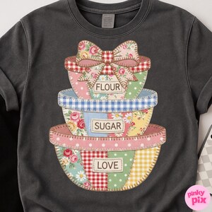 May include: Dark gray sweatshirt featuring a stack of patchwork bowls with the words "FLOUR", "SUGAR", and "LOVE". The bowls have floral, gingham, and polka dot patterns, topped with a large bow.