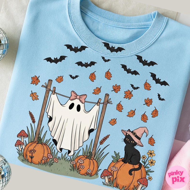 May include: Light blue sweatshirt with a Halloween-themed graphic. The design features a ghost with a pink bow, bats, pumpkins, fall leaves, a black cat wearing a witch hat, and mushrooms. The overall theme is a cute and whimsical Halloween design.