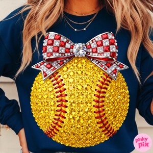 May include: Navy blue sweatshirt with a softball design. The softball is covered in yellow rhinestones with red stitching. A red and white checkered bow with rhinestone accents sits above the softball. A large, clear rhinestone is in the center of the bow.
