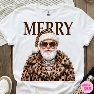 以下が含まれることがあります： 白い半袖Tシャツ。茶色の文字で「MERRY」と書かれ、その下にヒョウ柄のサンタ帽子、ヒョウ柄のコート、ヒョウ柄のサングラス、クリスタルのステートメントネックレスを身につけた白い髭の人物のグラフィックがプリントされています。