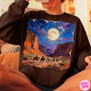 May include: Brown sweatshirt featuring a vibrant, artistic depiction of a desert landscape under a full moon. The artwork shows cowboys on horseback, riding through a canyon. The color palette includes blues, oranges, and browns.