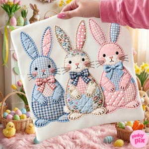 Op de afbeelding: Een witte stof met drie geappliqueerde paashazen. Elke konijn heeft een ander patroon, waaronder ruitjes, bloemen en stippen, in tinten blauw, roze en wit. De konijnen hebben strikjes en gestikte details.