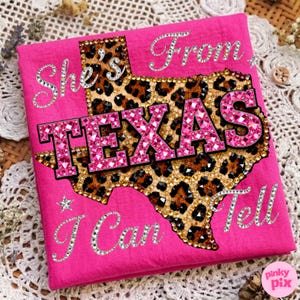 May include: A hot pink t-shirt with the text "She's From Texas I Can Tell". The Texas outline is filled with a leopard print design and embellished with rhinestones. The word "TEXAS" is in large, pink, rhinestone-covered letters.