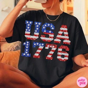 K&ouml;nnte beinhalten: Schwarzes T-Shirt mit dem Text "USA 1776" in Rot, Wei&szlig; und Blau. Die Buchstaben und Zahlen sind mit einem glitzernden Design gef&uuml;llt. Das T-Shirt hat einen l&auml;ssigen Stil.