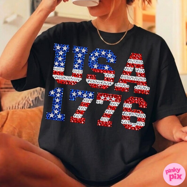 K&ouml;nnte beinhalten: Schwarzes T-Shirt mit dem Text "USA 1776" in Rot, Wei&szlig; und Blau. Die Buchstaben und Zahlen sind mit einem glitzernden Design gef&uuml;llt. Das T-Shirt hat einen l&auml;ssigen Stil.