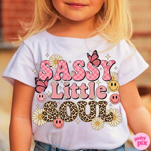 May include: White t-shirt with the text "SASSY Little SOUL" in pink, leopard print, and yellow. The design includes butterflies, daisies, and smiley faces. The shirt is a children's apparel item.